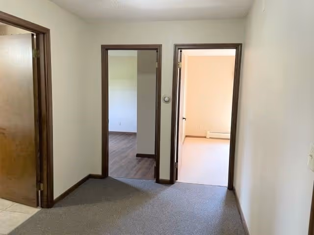 Interior view of a hallway in an assisted living facility with three doorways leading to different rooms. The walls are painted white, and the door frames are dark wood. The floor is carpeted in the hallway, with visible tile and wood flooring in adjacent rooms.