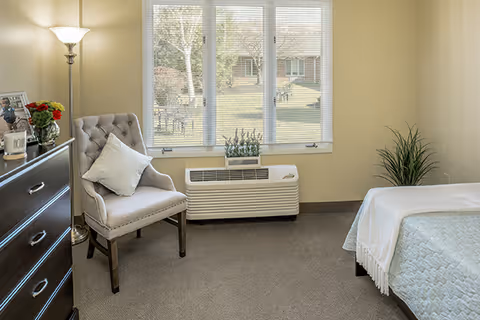 A cozy bedroom with a beige armchair with a pillow, a dark wooden dresser with framed photos and a vase of red flowers, a floor lamp, a bed with a white blanket, a potted plant, and a large window showing an outdoor view with trees and a building.