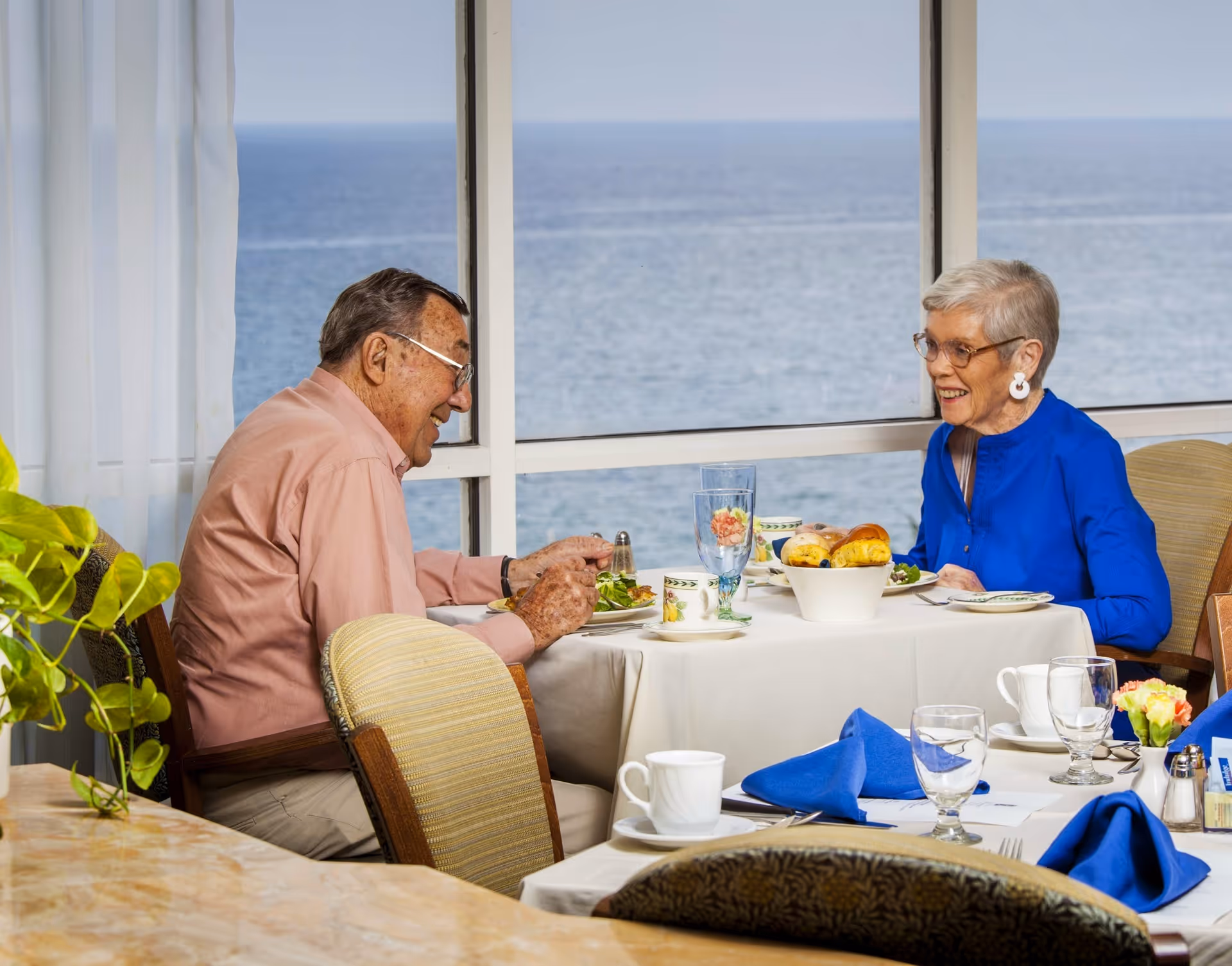 An elderly man and woman are sitting at a dining table by a large window with an ocean view. They are enjoying a meal together, with plates of food, glasses, and a basket of bread on the table. The woman is wearing a bright blue top and glasses, and the man is wearing a light pink shirt and glasses. The setting appears bright and cheerful with a plant visible on the left side.