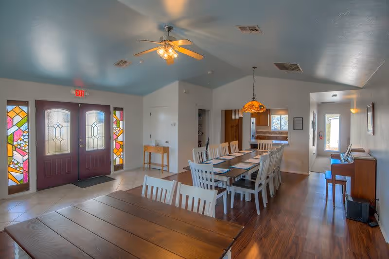Interior view of a dining room with two long wooden tables surrounded by white chairs. The room has a blue ceiling with a ceiling fan and a hanging stained glass lamp above one table. There are stained glass windows flanking a double door with an exit sign above it. A piano is positioned against the right wall near a hallway leading to another door.
