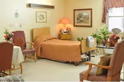 A nursing home bedroom with a single bed covered in a brown quilt, a bedside table with a lamp and flowers, two armchairs, a small table with a floral arrangement, a window with curtains, and framed artwork on the walls.
