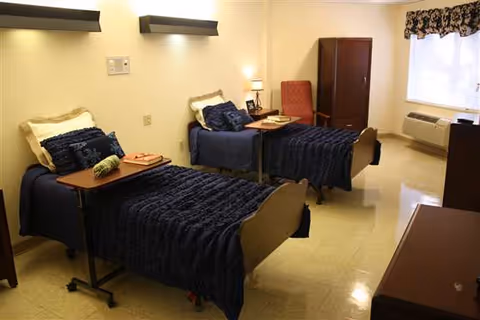 Shared nursing facility bedroom with two beds dressed in dark blue covers, overbed tables, a chair, dresser and a window air conditioner.