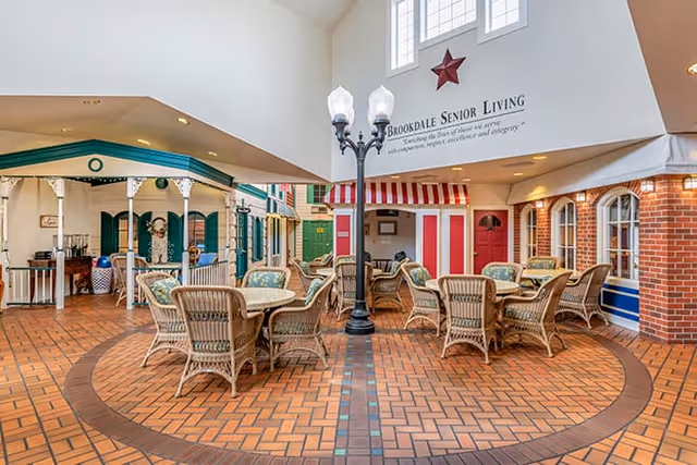 Indoor senior living community common area with wicker tables and chairs, storefront-style facades, and a central lamppost under a high ceiling.