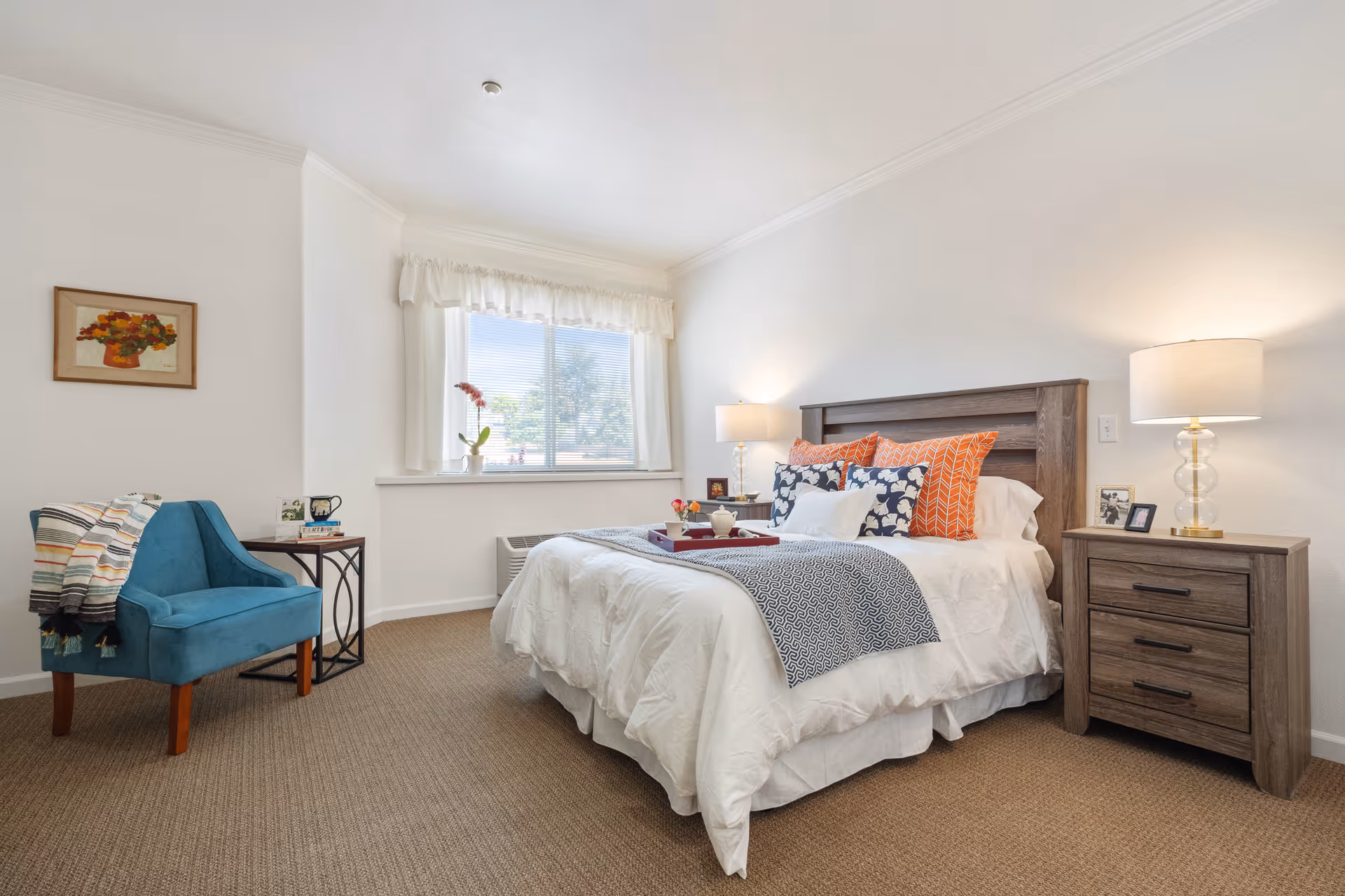 Bright furnished bedroom with a made bed, wooden nightstands and lamps, a blue accent chair, and a window with a vase on the sill.