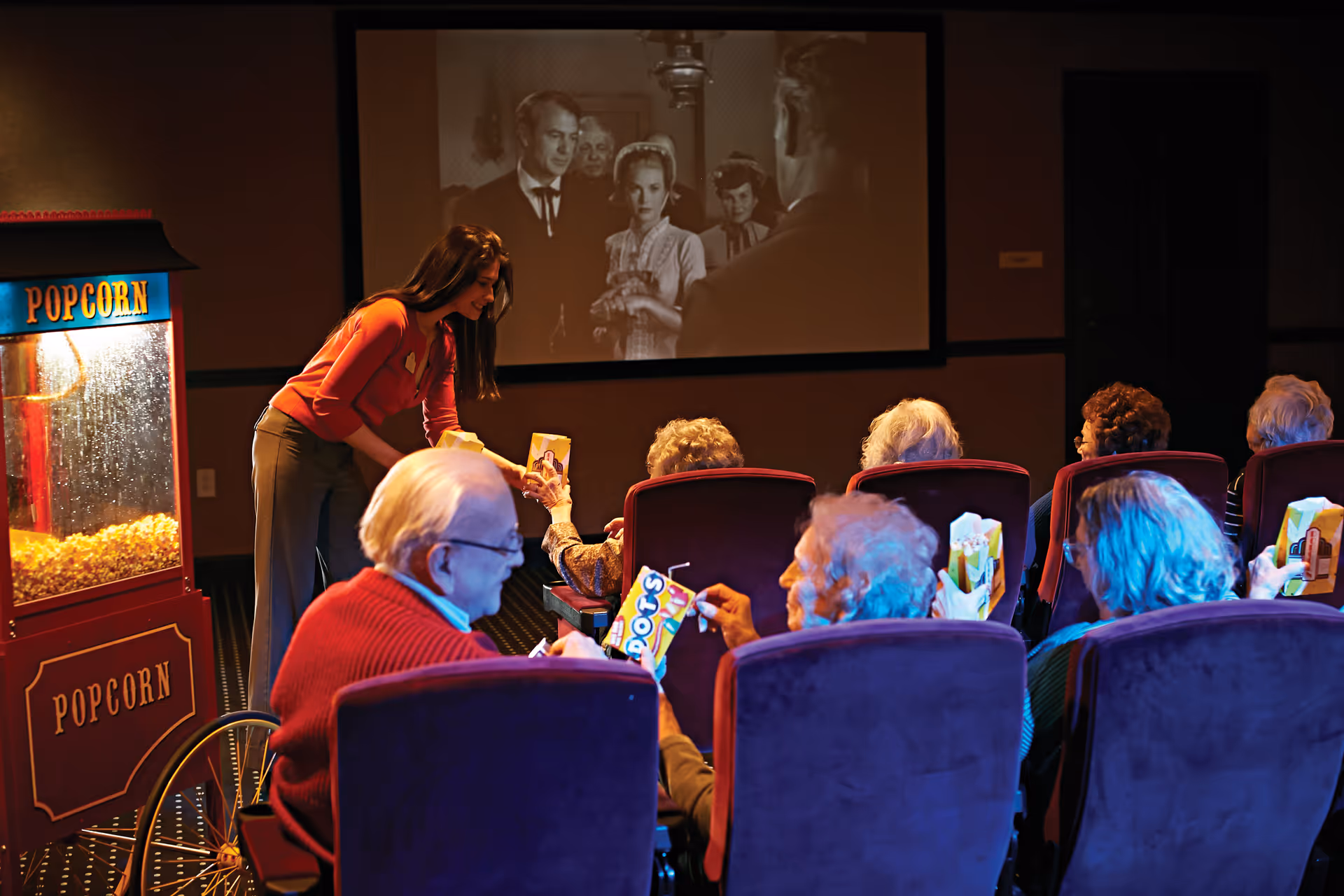 A group of elderly people seated in a small theater room watching a black-and-white movie on a large screen. A woman is handing out snacks to the seated audience. There is a popcorn machine on the left side of the image.