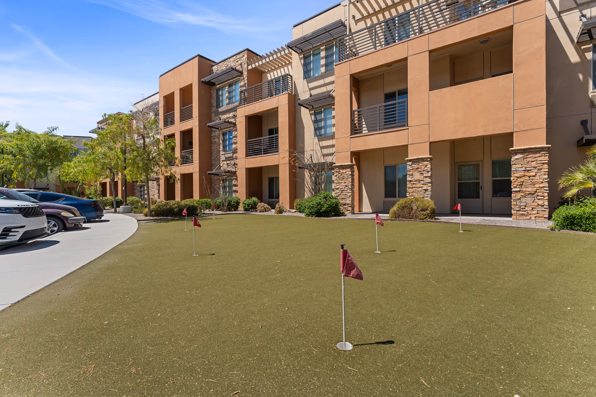 Outdoor putting green with several small red flags in front of a multi-story residential building with balconies and stone accents. Several parked cars are visible on the left side along a curved driveway, and there are trees and shrubs around the building.