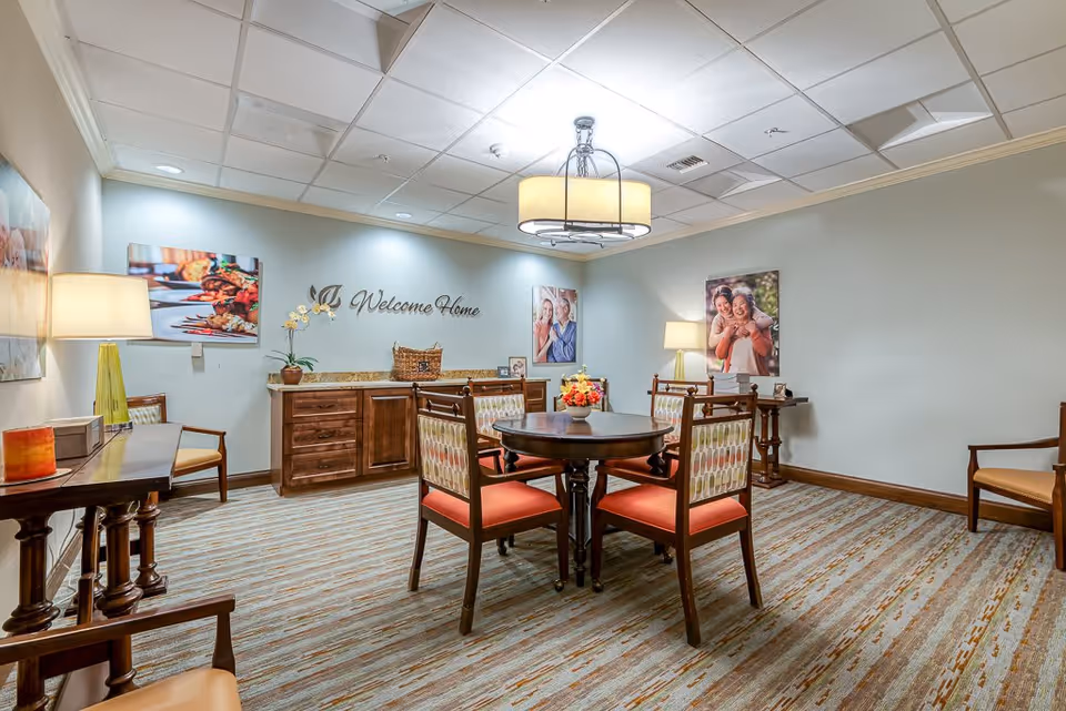 A warmly lit interior room with a round wooden table surrounded by four chairs with patterned backs and orange cushions. The room has a carpeted floor with a striped pattern and light-colored walls adorned with framed photos of smiling people and a decorative sign that reads 'Welcome Home'. There are two lamps on side tables and a ceiling light fixture above the table.