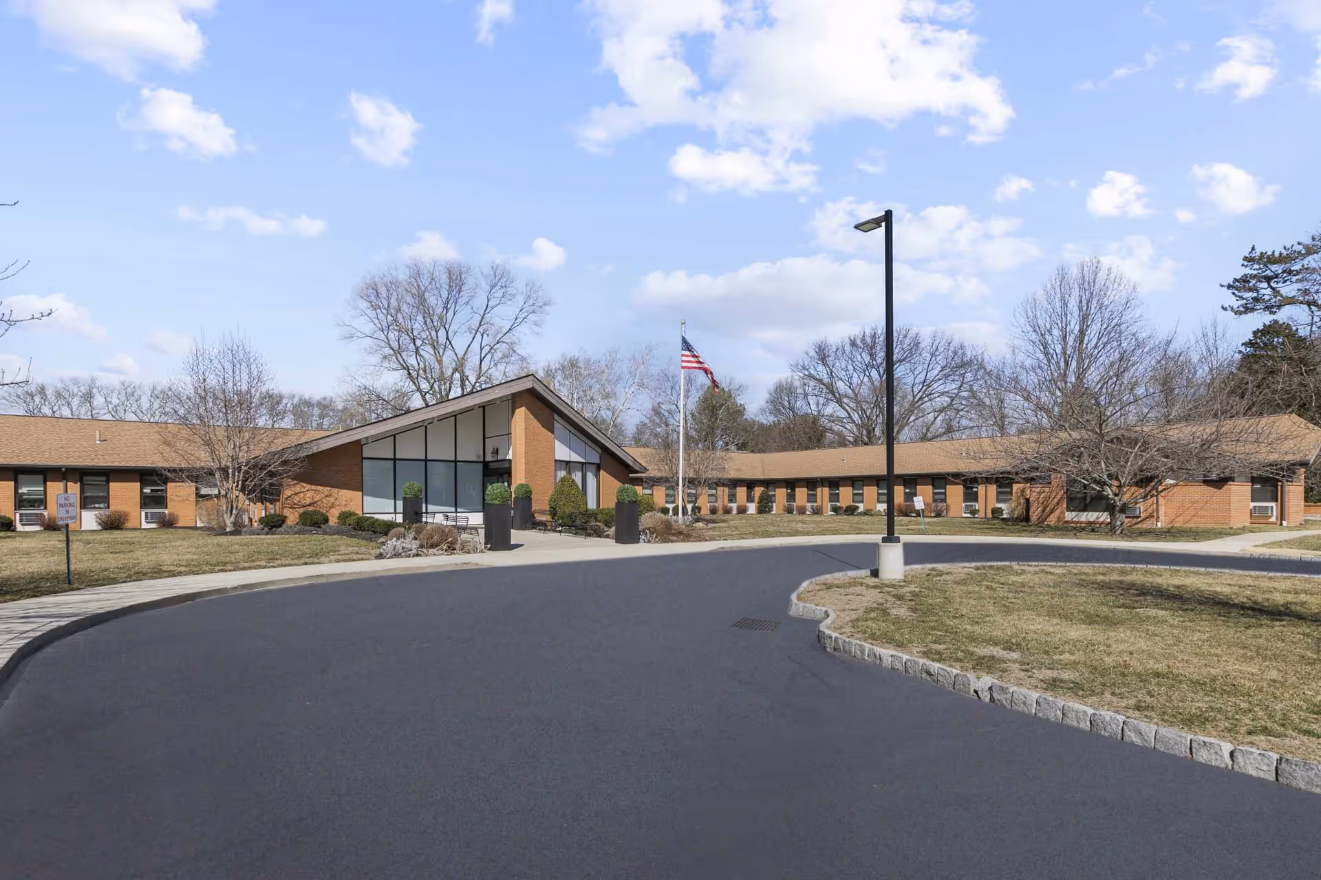 Exterior view of Rosewood Gardens Rehabilitation And Nursing Center showing a single-story brick building with large windows, an American flag on a flagpole, a curved driveway, and leafless trees under a partly cloudy sky.