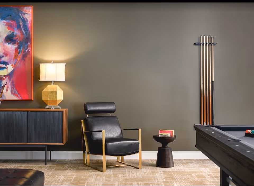 A modern lounge area featuring a black leather armchair with gold frame, a small round black side table with a book on it, a pool table with billiard balls, a wall-mounted rack holding pool cues, a wooden cabinet with a geometric gold lamp on top, and a colorful abstract portrait painting on the wall.