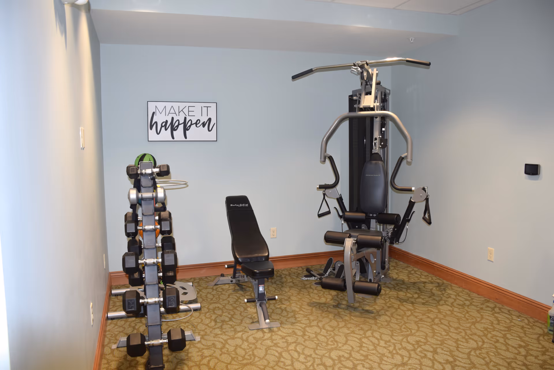 Small fitness room with a weight bench, a multi-function exercise machine, and a rack of dumbbells against a light blue wall. A framed sign on the wall reads 'MAKE IT happen'. The floor is carpeted with a green patterned carpet and the room has wooden baseboards.