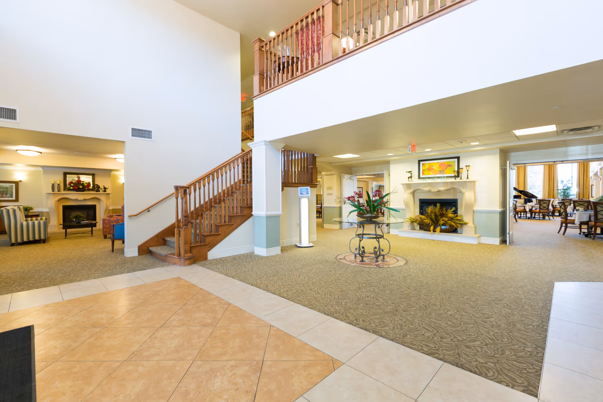 Spacious interior of Shawnee Hills facility featuring a tiled floor entrance leading to a carpeted area with a wooden staircase and a decorative table with flowers. There are two fireplaces with floral decorations, comfortable seating areas, and a dining room visible in the background with tables and chairs. The space is well-lit with ceiling lights and natural light from windows.