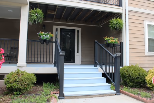 Front entrance of a senior living facility with a covered porch, stairs, railing, and hanging plants.
