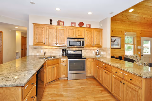 A spacious kitchen with wooden cabinets and granite countertops. The kitchen features a stainless steel oven with a microwave above it, a coffee maker, and a stand mixer. The floor is wooden, and the kitchen opens into a living area with wood-paneled walls and windows letting in natural light.