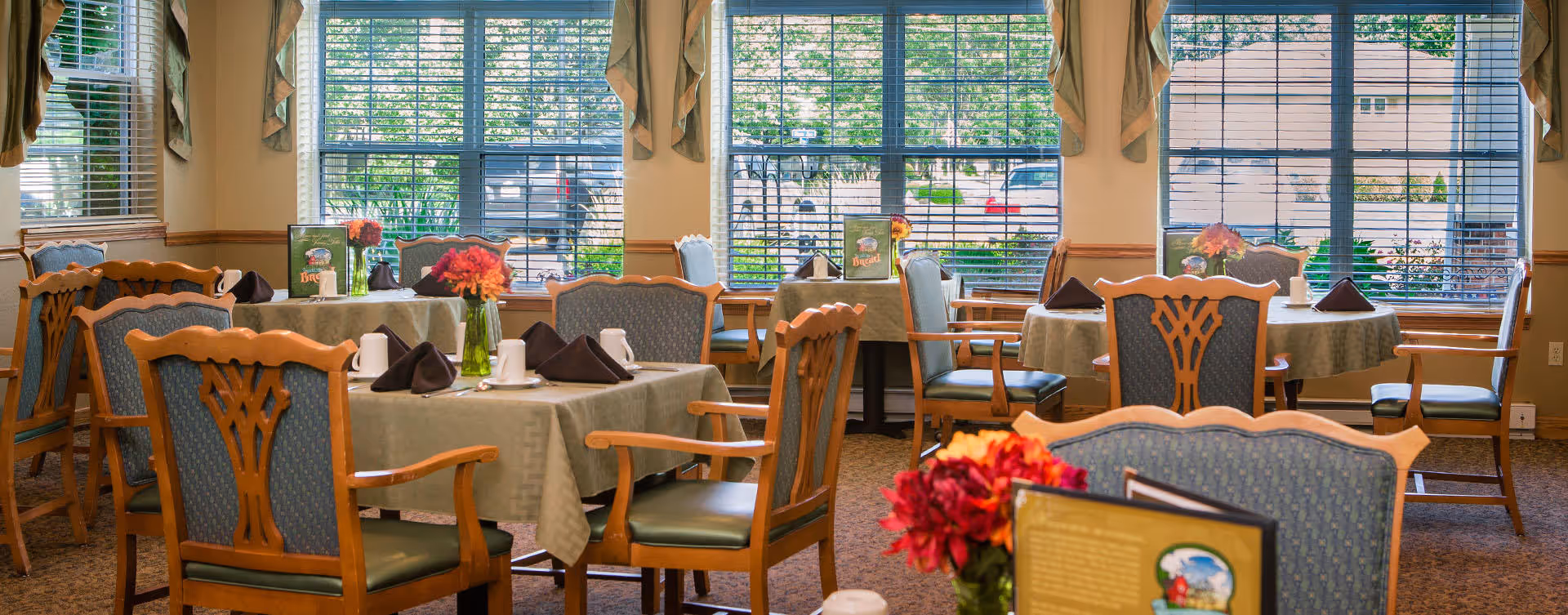 A bright dining room with several round tables covered with beige tablecloths, each set with white cups, dark folded napkins, and small flower vases. The room has large windows with blinds allowing natural light to fill the space, and wooden chairs with blue upholstered seats and backs surround the tables.