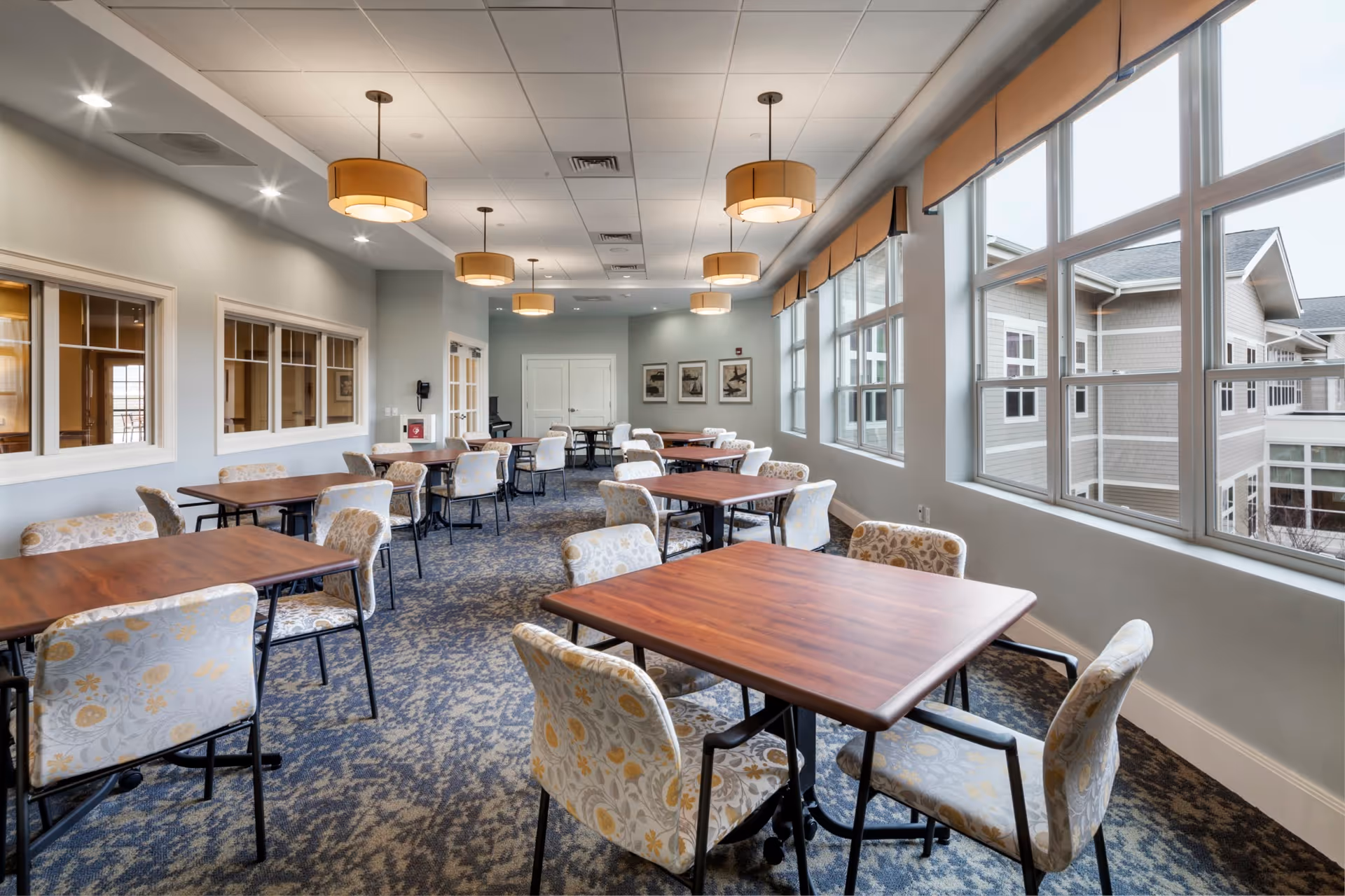 A bright dining room with multiple wooden tables and cushioned chairs arranged neatly. Large windows on one side let in natural light and offer a view of the exterior building. The ceiling has recessed lighting and several hanging pendant lights with beige shades. The walls are painted light gray, and there are framed pictures on the far wall.