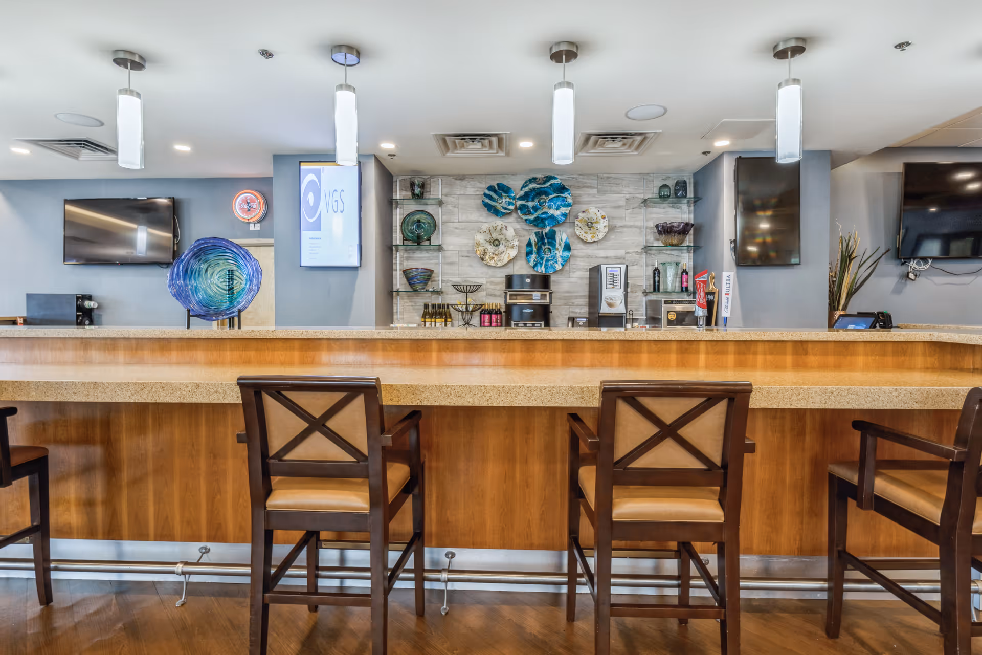 Interior view of a modern bar area with a wooden counter and four high chairs. Behind the counter, there are decorative glass plates mounted on the wall, shelves with glassware, and beverage dispensers. The space is well-lit with pendant lights hanging from the ceiling and multiple flat-screen TVs mounted on the walls.
