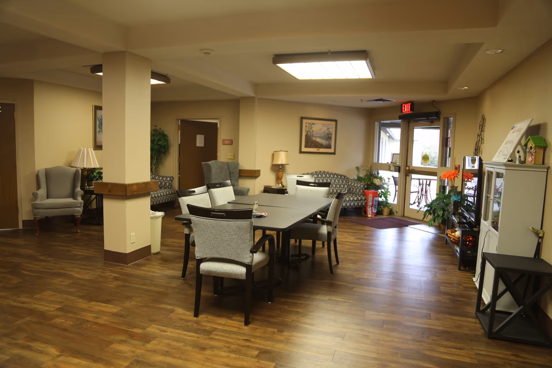 A common area in a senior living facility with wooden flooring, a rectangular table surrounded by six chairs, two armchairs, and two patterned sofas. The room has beige walls, a ceiling light, a lamp on a side table, framed artwork, plants, and a glass door leading outside with an exit sign above it.