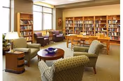 Sunlit community library lounge with bookshelves, upholstered chairs, tables, and lamps.