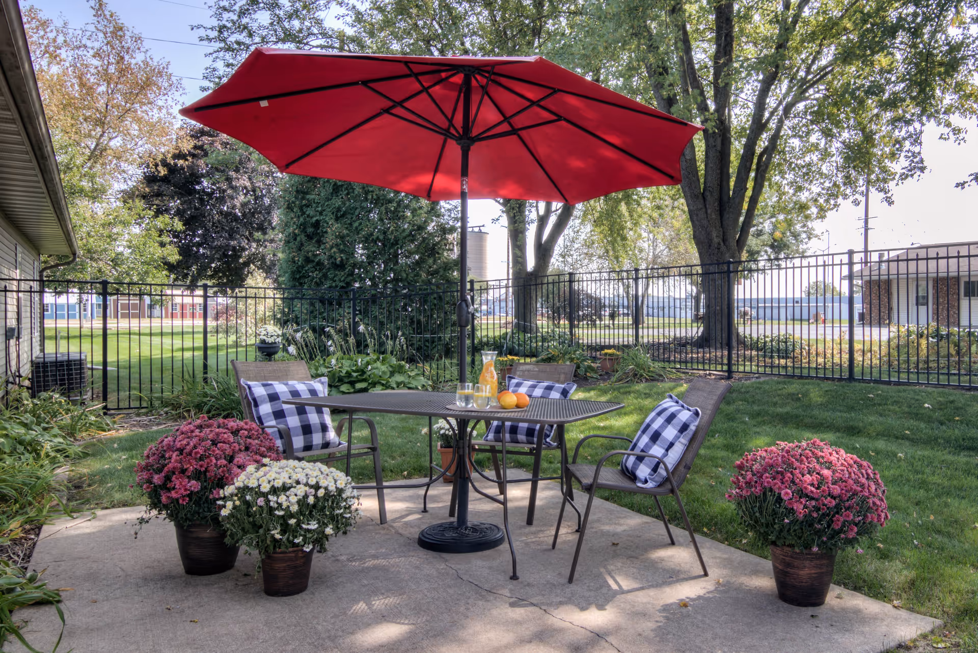 Outdoor patio with a table and four chairs under a red umbrella, surrounded by potted flowers and a fenced lawn.