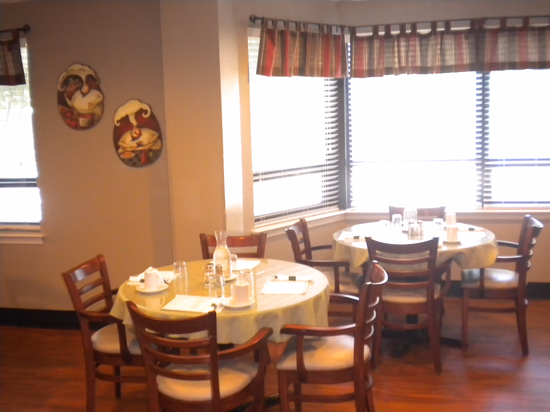 Two round dining tables set with white tablecloths, glasses, cups, and menus in a room with wooden chairs and large windows with blinds and plaid valances.
