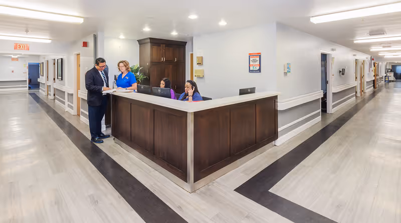 A reception desk area inside a senior living facility with two staff members seated behind the desk working on computers and a man and a woman standing in front of the desk reviewing documents. The hallway extends on both sides with light-colored walls and flooring, and handrails along the walls.