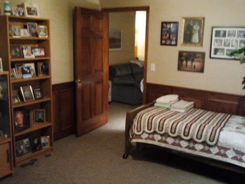 A cozy bedroom in a retirement home featuring a single bed with a patterned quilt and folded towels on top. The room has beige walls with wooden wainscoting and several framed pictures hanging. A wooden bookshelf filled with framed photographs is on the left side, and an open wooden door reveals a glimpse of a living room with a sofa.
