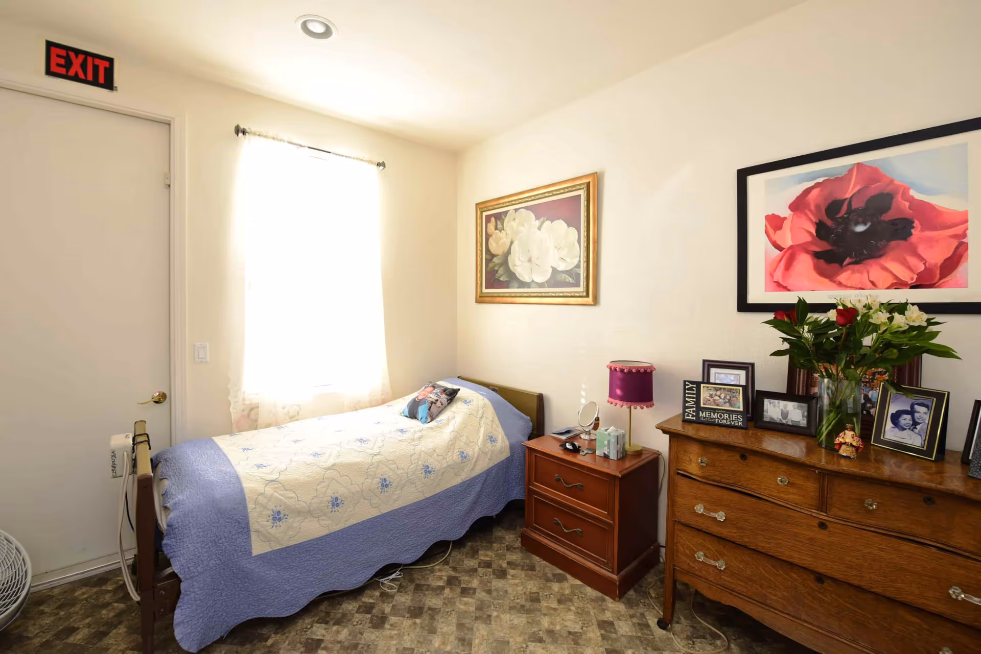 A small, cozy bedroom with a single bed covered in a light blue and cream quilt. There is a wooden nightstand with a purple lamp and some personal items next to the bed. A wooden dresser with framed photos, a vase of flowers, and decorative items is against the opposite wall. Two framed floral paintings hang on the walls. A window with sheer curtains allows natural light to brighten the room. An exit sign is above a closed door.