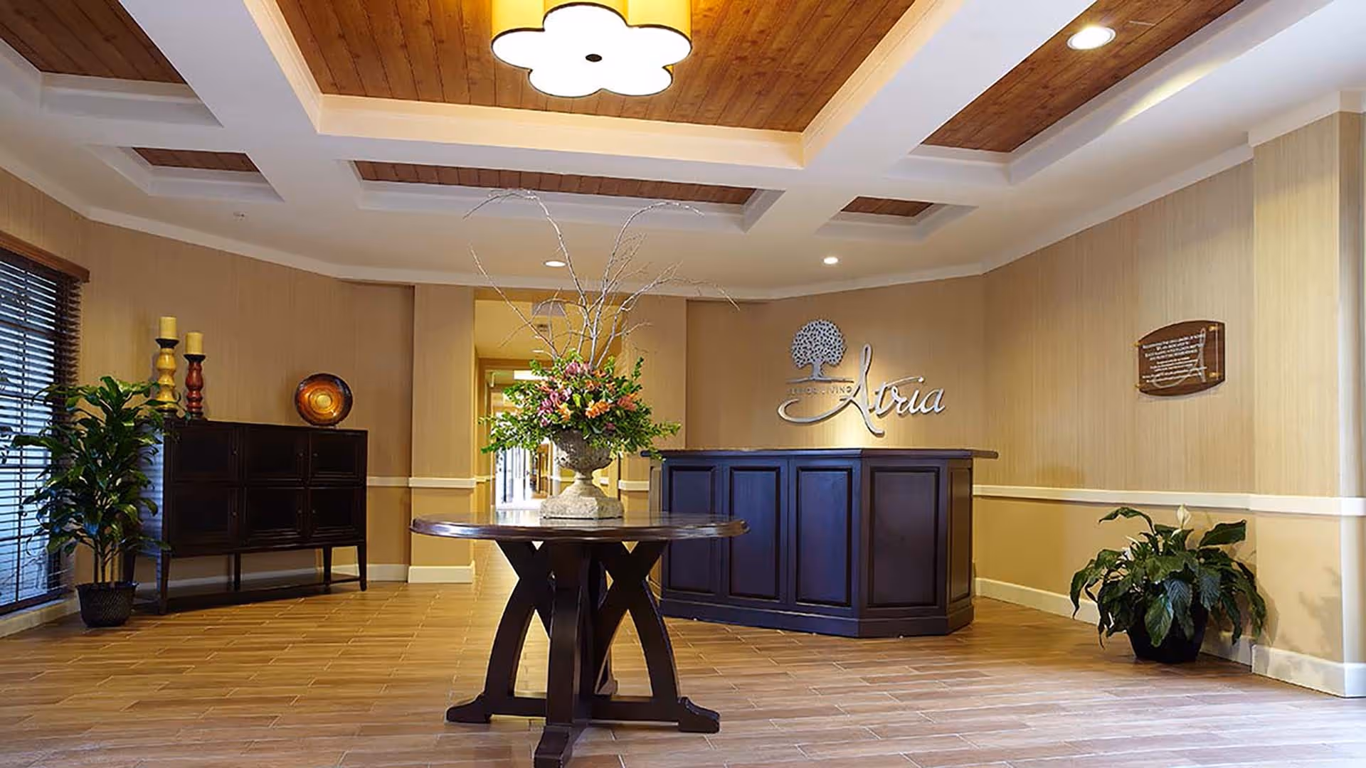 Reception lobby with a round table holding a floral arrangement and a dark wood front desk beneath a coffered ceiling.
