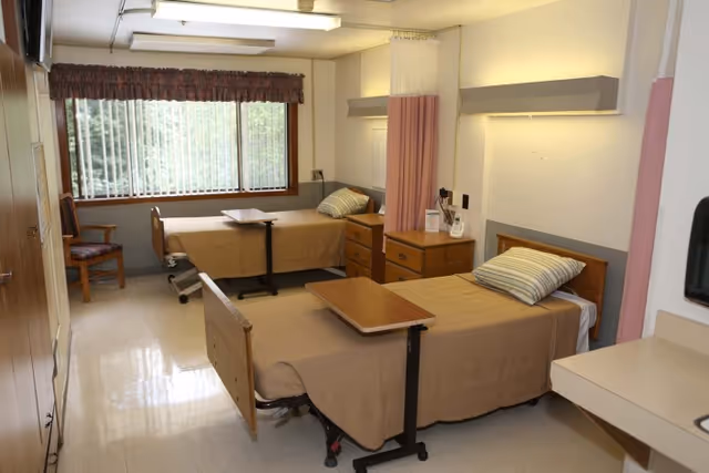 A rehabilitation facility room with two hospital-style beds, each with a beige blanket and a pillow. Each bed has an overbed table. There is a wooden dresser between the beds and a window with vertical blinds and a valance letting in natural light. A wooden chair is placed near the window. The room has a clean, clinical appearance with light-colored walls and flooring.