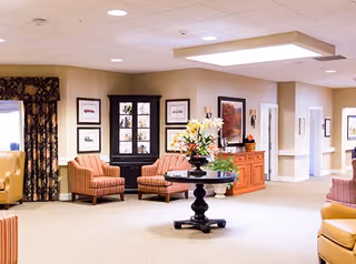 Well-lit senior living common lounge with upholstered chairs, a central round table with a floral arrangement, framed artwork and a hallway.