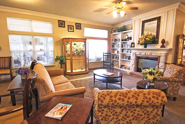 A cozy living room with a beige sofa, patterned armchairs, a round coffee table, and a fireplace with a large mirror above it. There are built-in bookshelves filled with books and decorative items, a glass display cabinet, and windows with blinds letting in natural light. The room has warm lighting from a ceiling fan with lights.