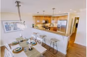 Bright and modern open-concept kitchen and dining area with wooden floors. The kitchen features wooden cabinets, stainless steel appliances including a refrigerator and microwave, and a granite countertop with three bar stools. The dining table is set with plates, bowls, glasses, and cutlery, and is illuminated by a hanging light fixture. A framed picture hangs on the wall near the dining area.