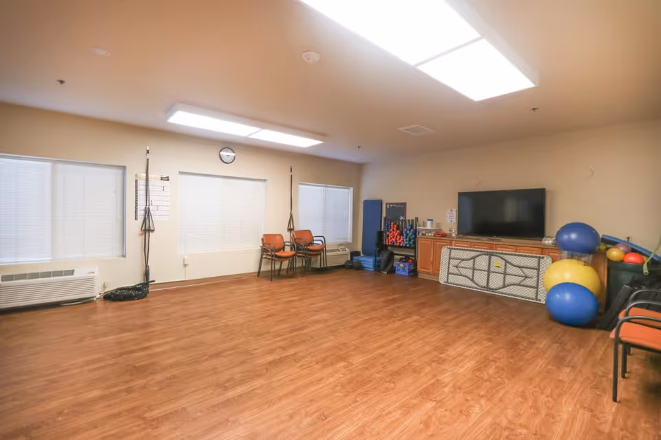 A spacious room with wooden flooring and beige walls, featuring three windows with closed blinds, a wall clock, several orange chairs stacked in the corner, exercise equipment including large colorful exercise balls, weights, and resistance bands, and a large flat-screen TV mounted above a cabinet.
