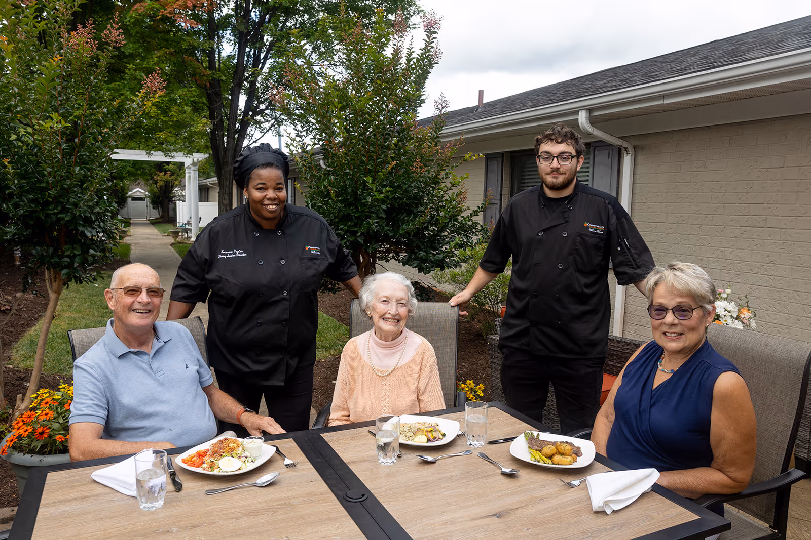 Two elderly women and one elderly man seated at an outdoor dining table with plates of food and glasses of water. Two staff members in black chef uniforms stand behind them, smiling. The setting is a garden area with trees, flowers, and a building in the background.