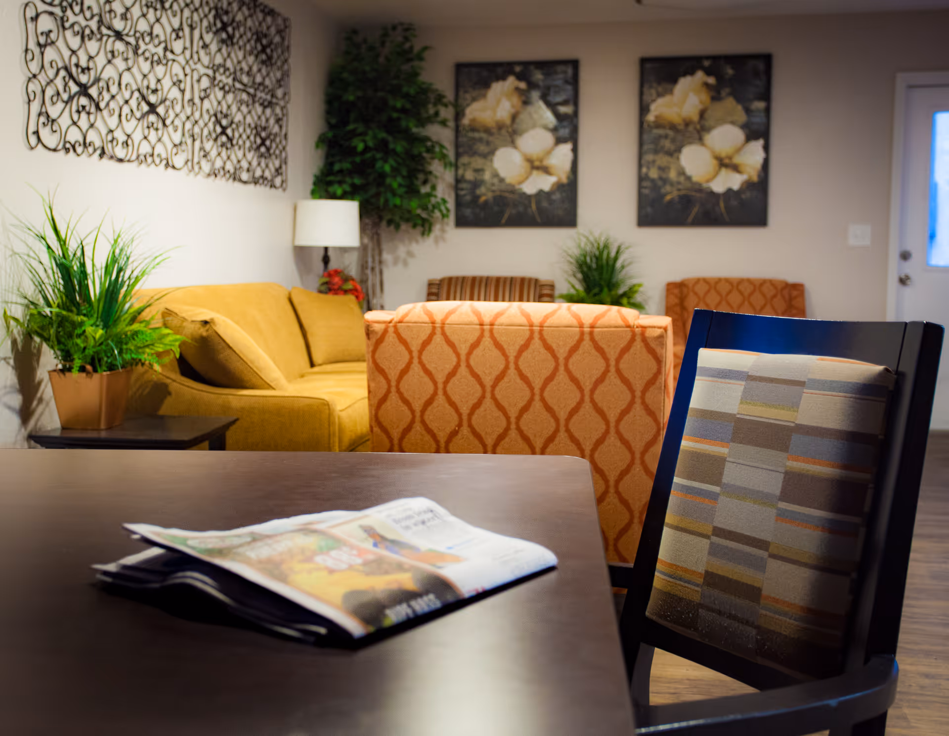 A cozy living room area with a dark wooden table in the foreground holding a folded newspaper. Behind the table, there is a chair with a striped cushion. Further back, there are two patterned armchairs, a mustard yellow sofa, a side table with a potted plant, a floor lamp, and two framed floral paintings on the wall. The room has a warm and inviting atmosphere with additional greenery and soft lighting.