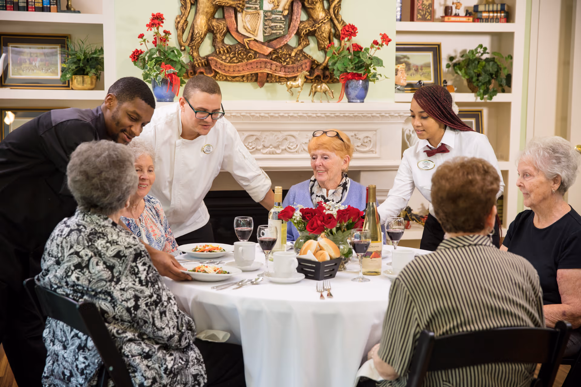 Several elderly residents seated around a round dining table with waitstaff serving food in a decorated communal dining room.