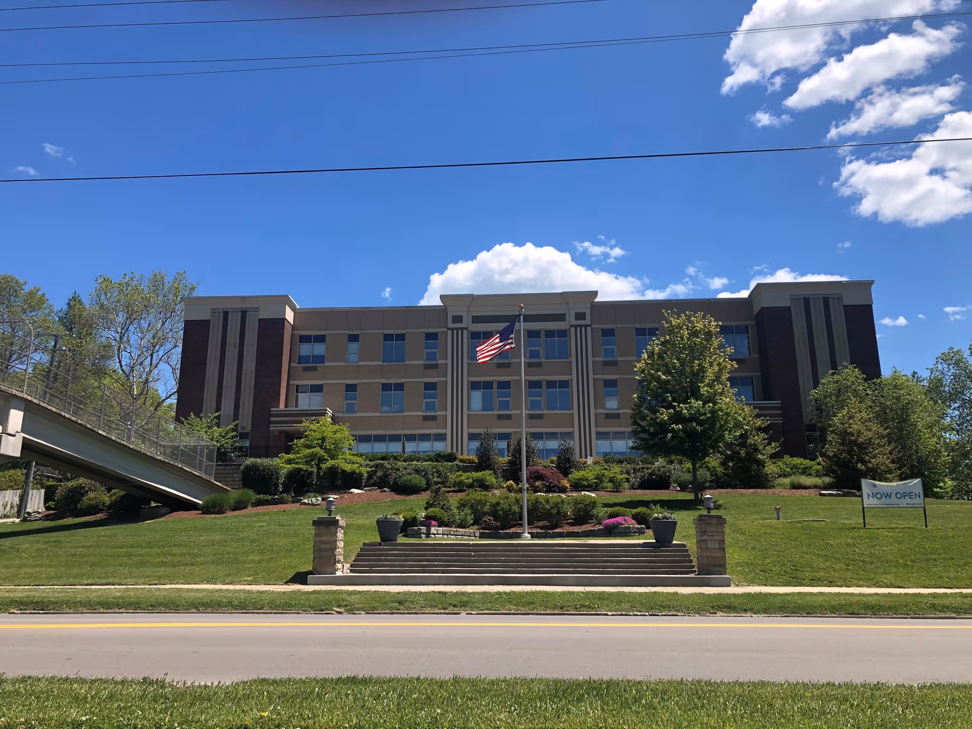 Front exterior view of a multi-story assisted living facility building with an American flag on a flagpole in the center of a landscaped lawn. There are stairs leading up to the building, trees, shrubs, and a sign on the right side that reads 'NOW OPEN'. The sky is clear with a few clouds.