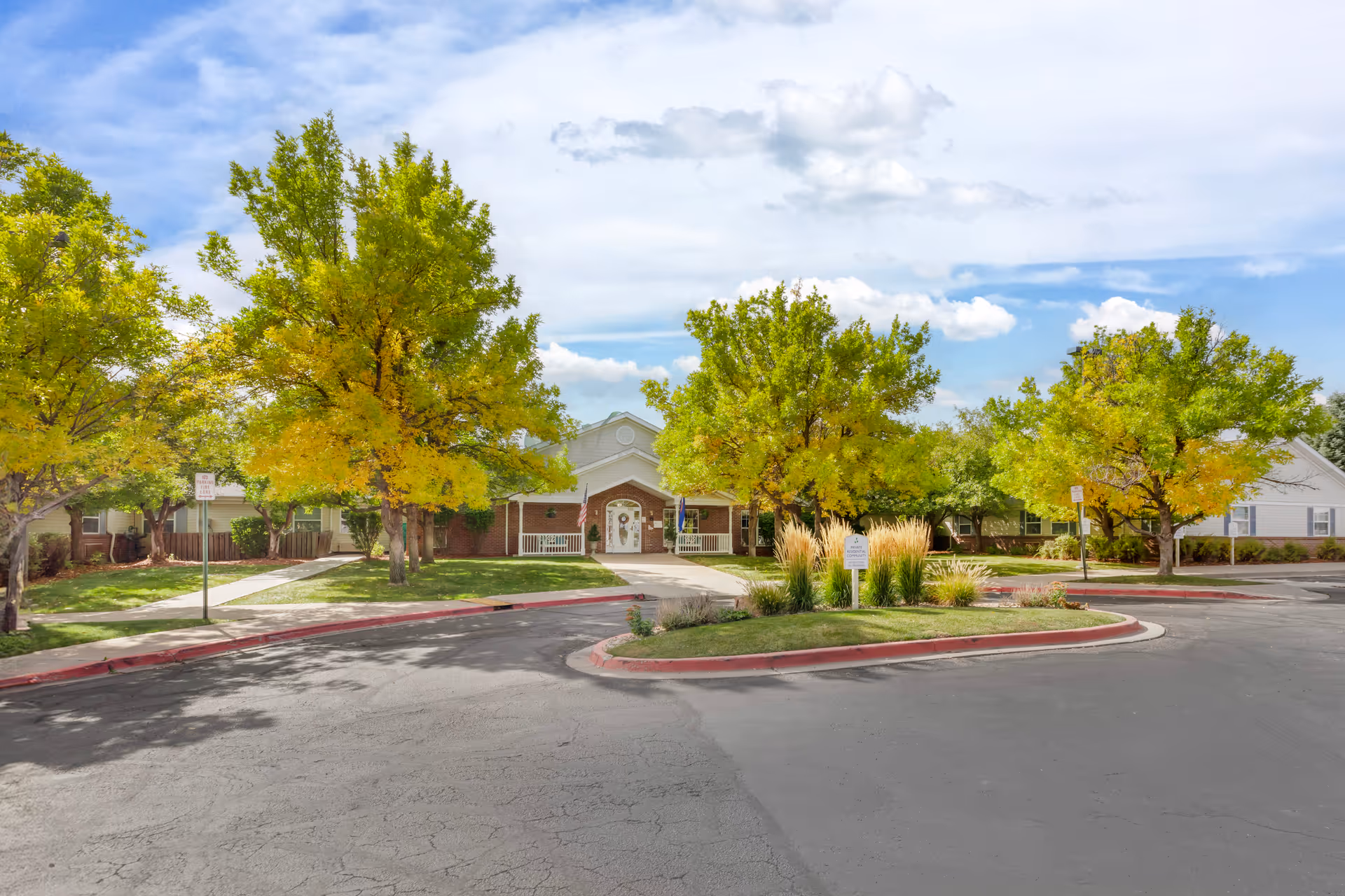 Front exterior view of Brookdale Pinehurst Park facility with a driveway, green trees with some yellow leaves, a lawn, and a building entrance with two white benches and flags.