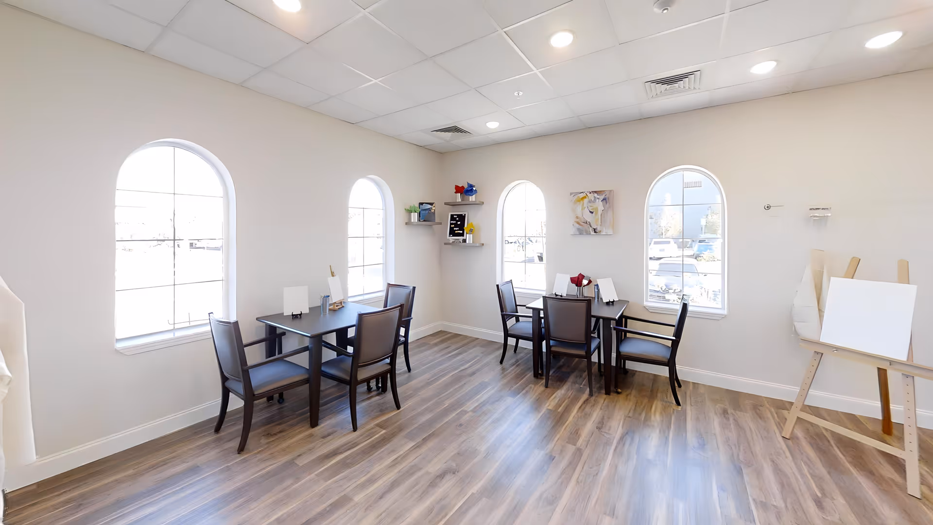 A bright room with four arched windows letting in natural light, two small dark wood tables each surrounded by four chairs, light-colored walls, wood flooring, a small wall shelf with decorative items, a painting on the wall, and an easel with a blank canvas.