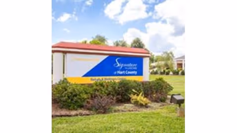 Outdoor view of a sign for Signature HealthCARE of Hart County Rehab & Wellness Center surrounded by green bushes and grass under a partly cloudy sky.