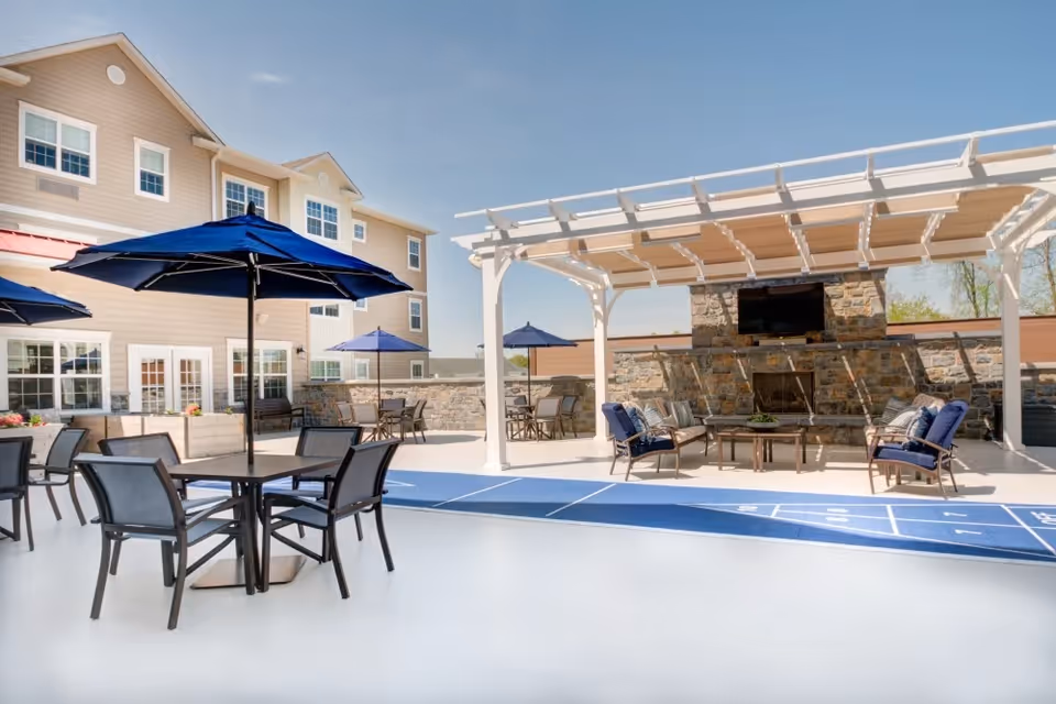 Outdoor patio area at Merrill Gardens at Glen Mills featuring multiple tables with blue umbrellas, chairs, a shuffleboard court, and a covered seating area with cushioned chairs and a stone fireplace with a mounted TV.