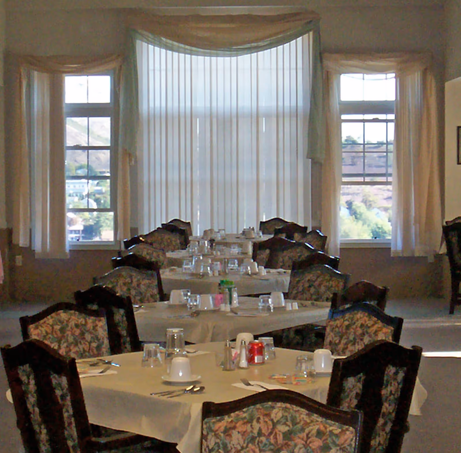 Dining room with multiple tables covered with white tablecloths, set with cups, glasses, utensils, and condiments. The room has large windows with sheer curtains allowing natural light to enter.