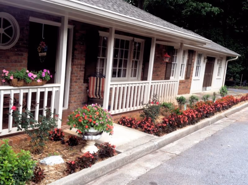 Exterior view of a senior care center building with a brick facade, white railings, and multiple doors and windows. There are flower beds with red and pink flowers along the sidewalk in front of the building.
