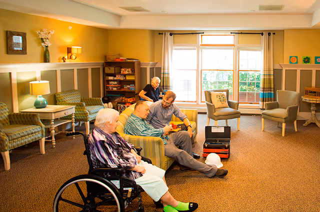 A bright senior living lounge with residents seated on sofas and chairs, including a woman in a wheelchair in the foreground.