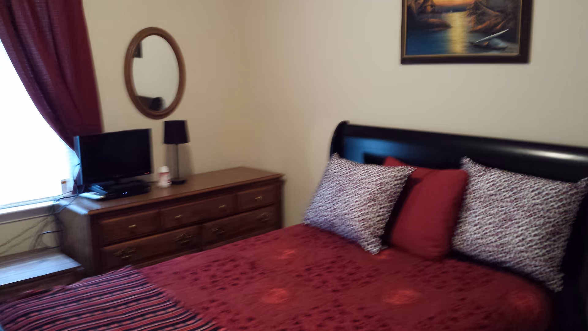 A bedroom with a bed covered in a red bedspread and three pillows, two patterned and one solid red. There is a wooden dresser with a small TV, a lamp, and a round mirror on the wall above it. A window with dark red curtains is partially visible on the left side, and a framed painting hangs on the wall above the bed.