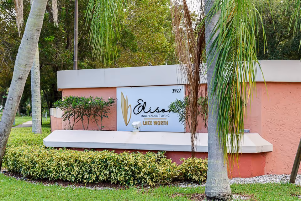 Outdoor view of the entrance sign for Elison Independent Living of Lake Worth, surrounded by palm trees and greenery.