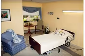 A cozy bedroom in a senior living facility featuring a hospital-style bed with white bedding and decorative pillows, a blue upholstered armchair with a blanket draped over it, a small round wooden table with two chairs near a window with blue valance curtains, and framed artwork on the wall.