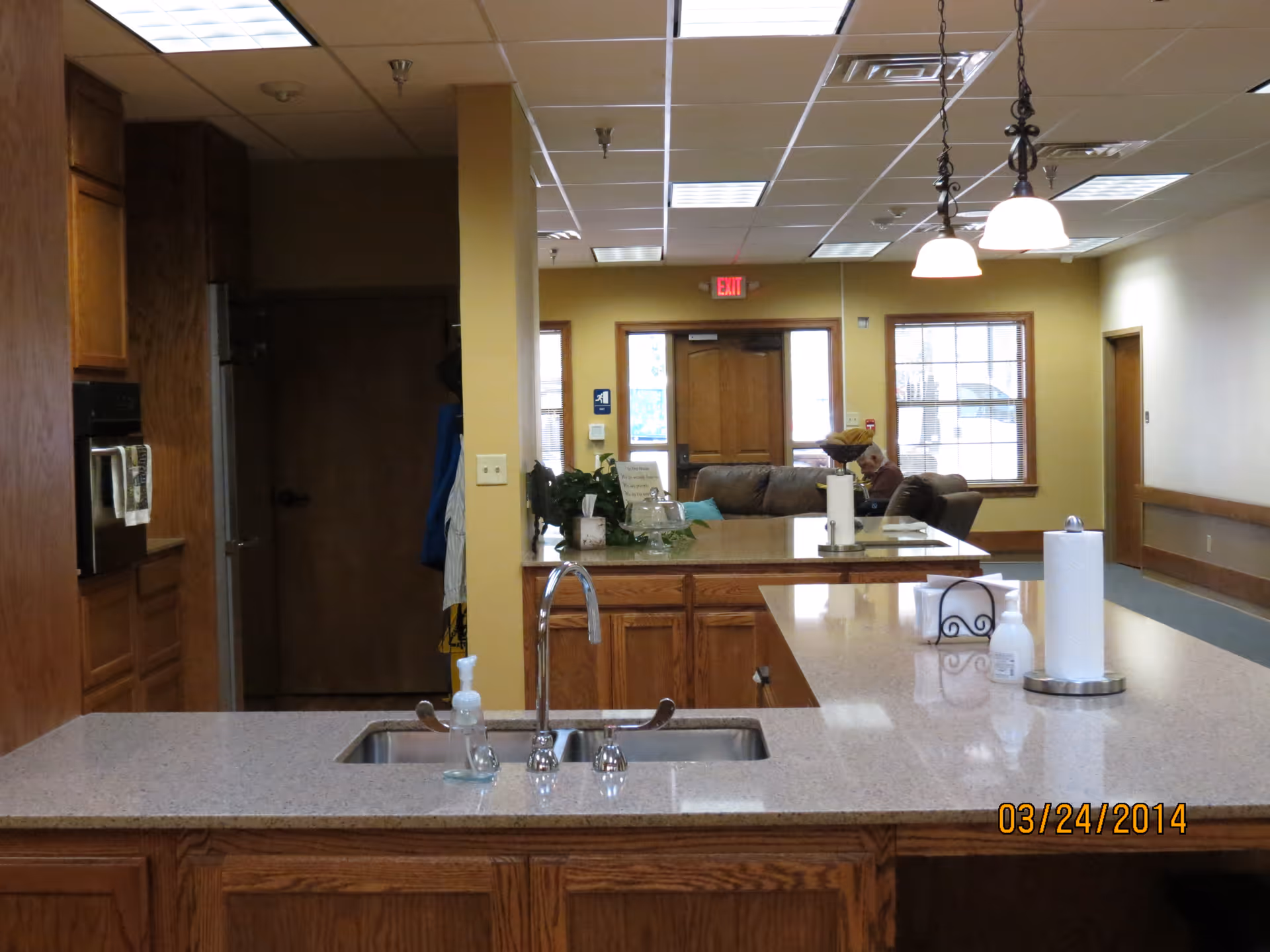 Interior view of a nursing center kitchen area with a countertop and sink in the foreground. The countertop has a soap dispenser and paper towel holder. In the background, there is a living room area with a couch and a person sitting on it near windows and a door. The ceiling has fluorescent lights and hanging pendant lights.