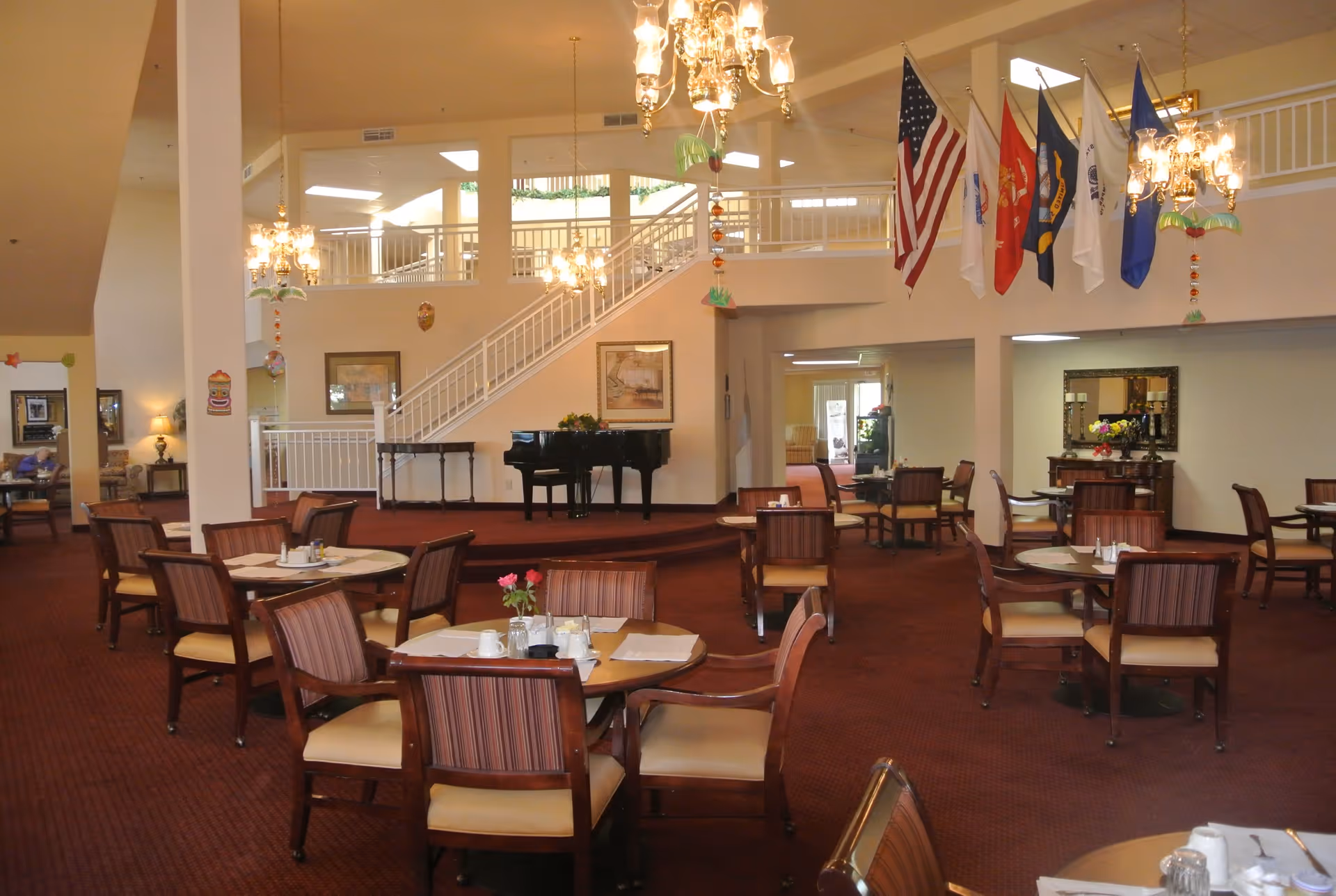 A spacious dining area with round tables and wooden chairs arranged neatly on a carpeted floor. The room features chandeliers hanging from the ceiling, several flags displayed on the upper wall, and a black grand piano on a small stage near a staircase. The walls are decorated with framed artwork and mirrors, and there are floral centerpieces on the tables.