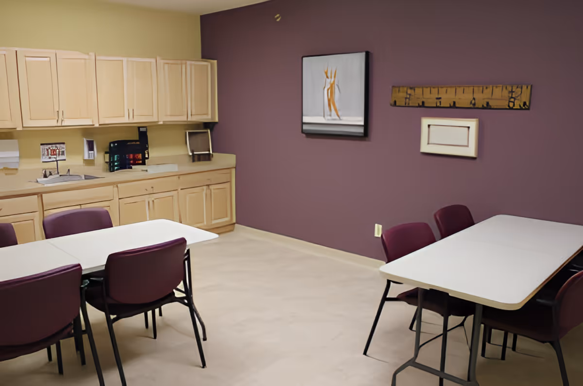 A room with beige cabinets along one wall, a sink, and two white tables surrounded by purple chairs. The walls are painted purple and beige, and there are three pieces of abstract artwork hanging on the purple wall.
